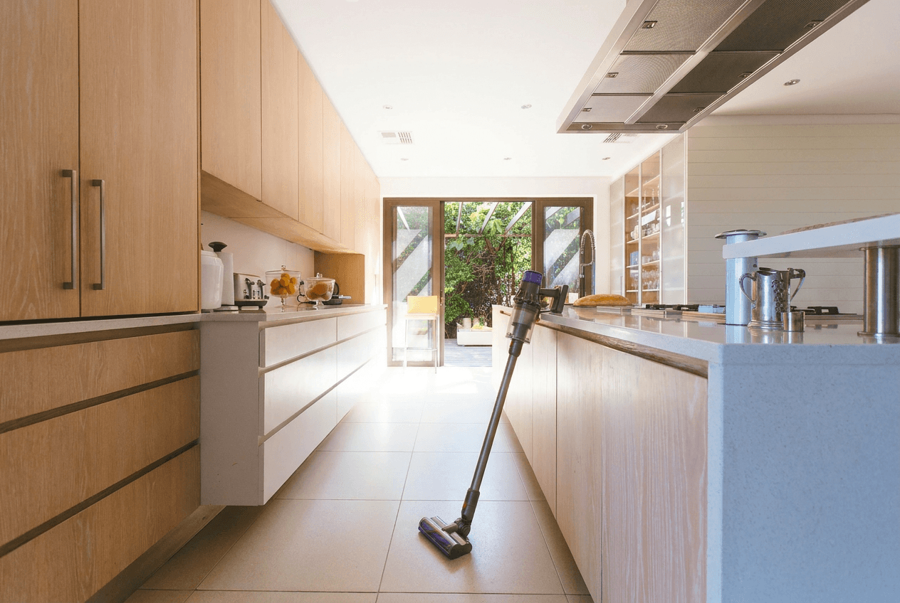 A modern, bright kitchen features light wood cabinets, white countertops, a tiled floor, and a Dyson stick vacuum leaning against the kitchen island.